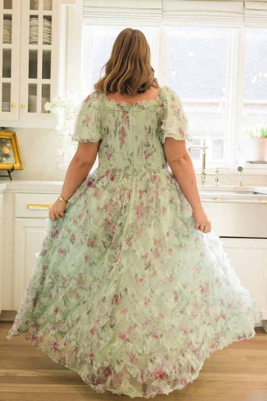 In a sunlit kitchen, a woman stands with her back turned, wearing the Ivy City Co Florentina Dress in Pistachio Aquarelle Fleur—a flowing chiffon dress adorned with ruffled sleeves. She lightly lifts the sides of her dress, highlighting its airy volume and delicate floral texture—an elegant maternity-friendly piece designed to celebrate every stage of motherhood.