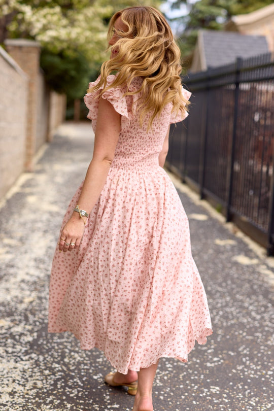 Wearing the Hattie Dress in Madeline Pink by Ivy City Co, a woman with wavy blonde hair strolls down a tree-lined path, glancing back over her shoulder.