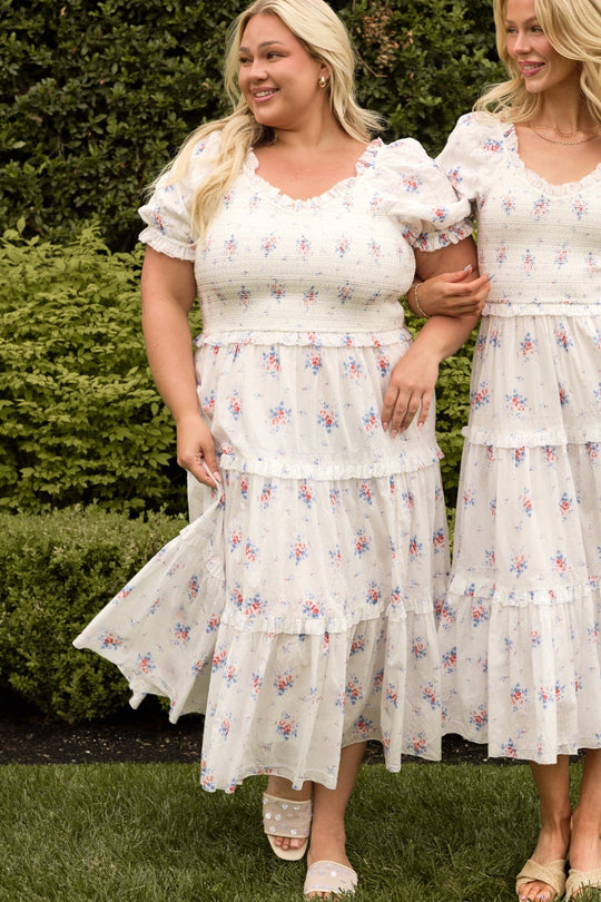 Two women stand together outdoors on the grass in front of green bushes, both smiling and wearing white, short-sleeved dresses with a delicate floral pattern. One woman holds the skirt of her Ivy City Co Emily Dress in Heritage Soft Ditsy Floral, a midi style designed to be nursing-friendly as well as perfect for maternity wear.