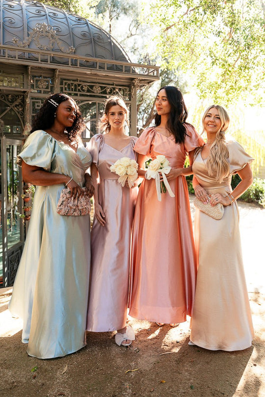 Four women wearing the Ivy City Co Tessie Dress in Champagne stand outdoors near a metal gazebo, each holding small bouquets and smiling, with sunlight filtering through the trees in the background.