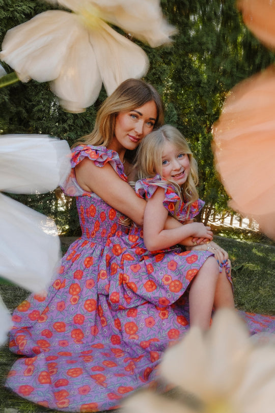 A woman and young girl wearing matching Ivy City Co Mini Hattie Dresses in the Thistle Flower Poppy Woodblock print sit on the grass, hugging and smiling, surrounded by blurred white and peach flowers with green trees in the background.