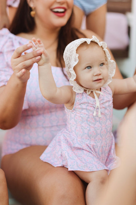 A smiling woman holds hands with a baby wearing the Baby Bitsy Swim Dress in Raquel Pink Floral and a frilly white bonnet. The baby looks up with raised arms as they share a joyful moment together.