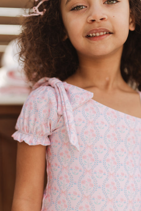 A young girl with curly hair smiles outdoors in soft sunlight, wearing the Ivy City Co Mini Sailor One Piece in Raquel Pink Floral.