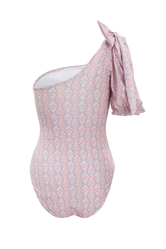 The Sailor One Piece in Raquel Pink Floral by Ivy City Co features a pink and light blue floral pattern, an asymmetrical neckline, and a large ruffled bow on the right shoulder, shown on a white background.