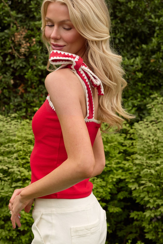 A woman with long blonde hair smiles outdoors in the Ivy City Co Sammi Top in Firework Red, featuring a white and red bow on the shoulder, paired with white pants—a chic Fourth of July look.
