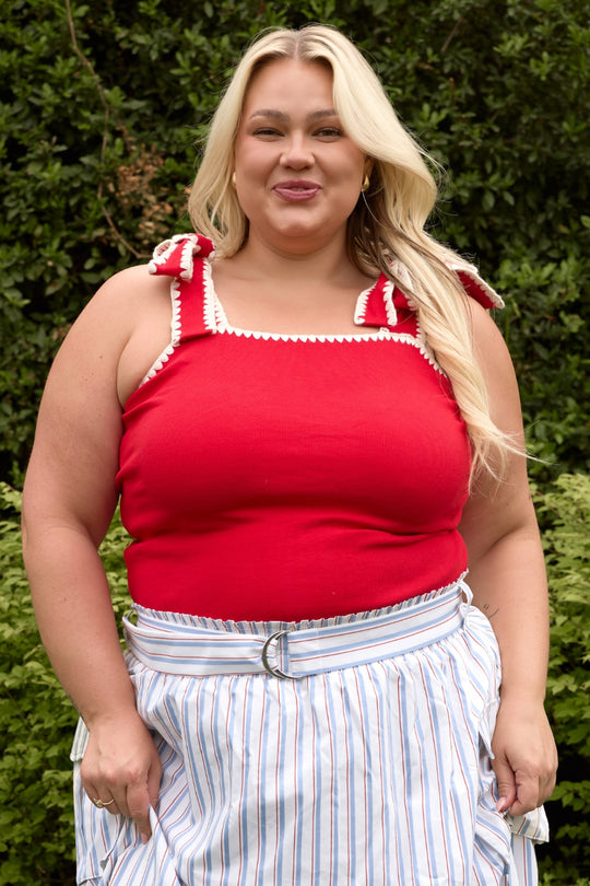 A woman with long blonde hair stands outdoors, smiling. She wears the Ivy City Co Sammi Top in Firework Red with a striped skirt and belt—a chic, maternity-friendly Fourth of July look set among lush green bushes.