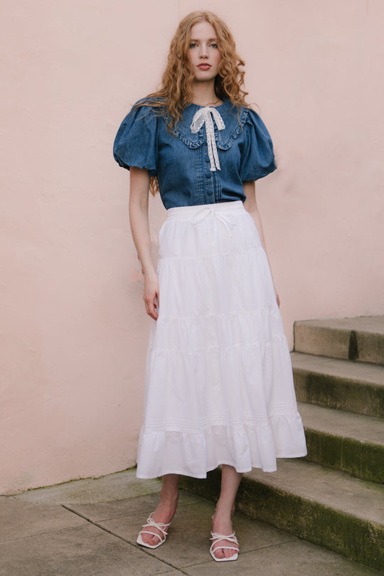 A woman with long, wavy red hair stands by the steps against a pale pink wall. She wears a blue puff-sleeve blouse, white sandals, and the Ivy City Co Sansa Skirt in White, an ankle-length cotton voile skirt with a lace bow.