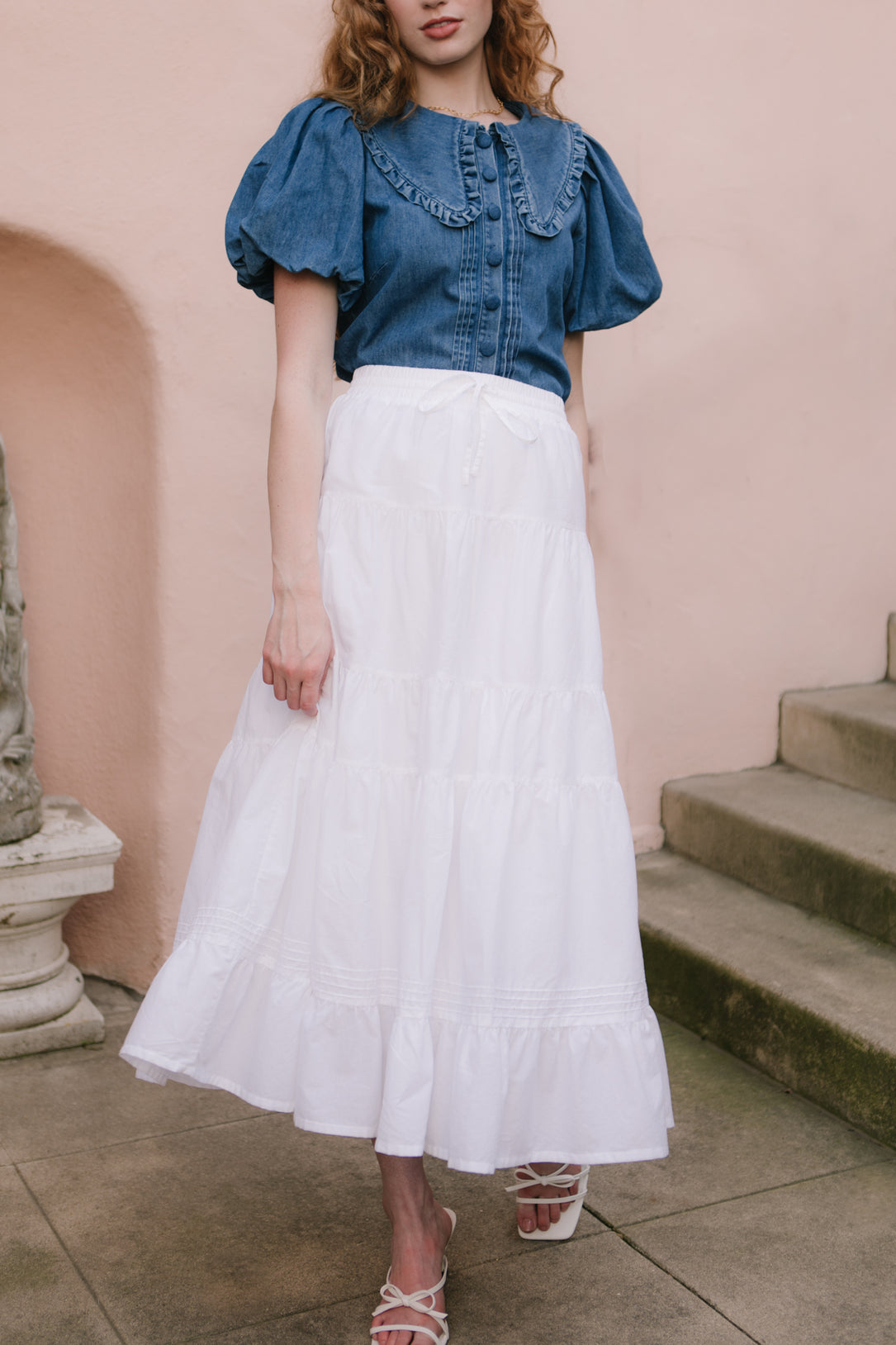 A woman stands outdoors wearing Ivy City Cos Sansa Skirt in White with a blue puff-sleeve blouse and white strappy sandals; a stone staircase and pink wall are seen in the background.