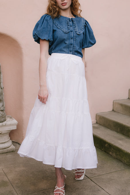 A woman stands outdoors wearing Ivy City Cos Sansa Skirt in White with a blue puff-sleeve blouse and white strappy sandals; a stone staircase and pink wall are seen in the background.