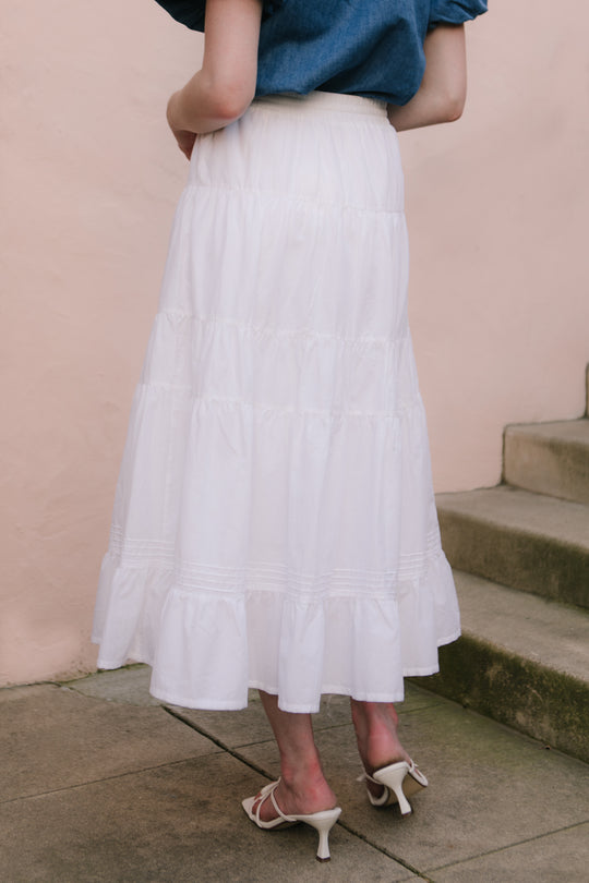 A person stands outdoors on concrete steps by a pale pink wall, wearing Ivy City Co’s Sansa Skirt in White—an ankle-length, maternity-friendly cotton voile skirt—paired with a blue top and white heeled sandals.
