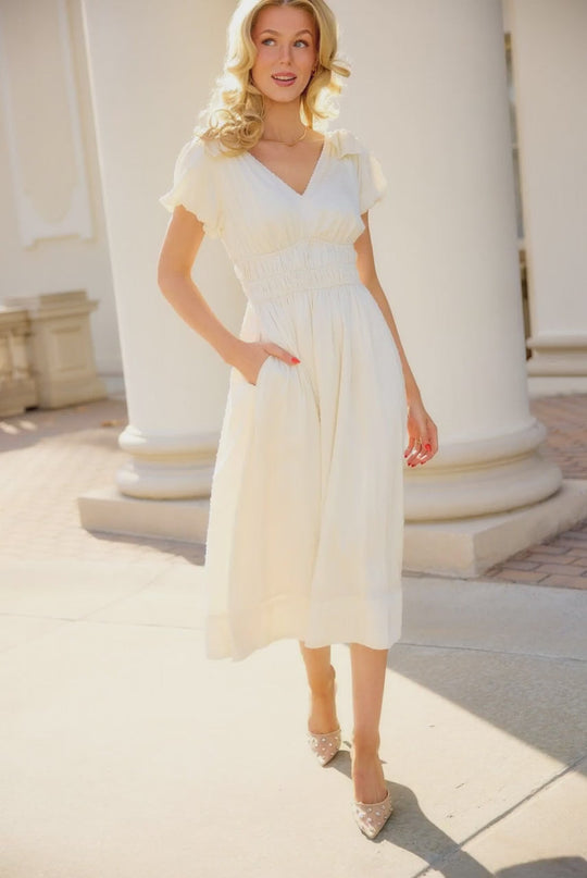 A woman, smiling and looking slightly away from the camera, stands outdoors near tall white columns in the Zella Bow Dress in Ivory by Ivy City Co. The elegant midi dress features short sleeves and convenient pockets, while sunlight casts soft shadows on the ground, highlighting its timeless style—making it a perfect choice for a rehearsal dinner.