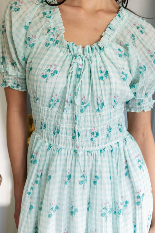A person wearing the Ivy City Co Jessie Dress in Bloom Gingham—a light blue, short-sleeved dress featuring a floral print, ruching at the waist and neckline, and delicate ruffle details. This soft, summery style is designed to be both nursing friendly and maternity friendly.