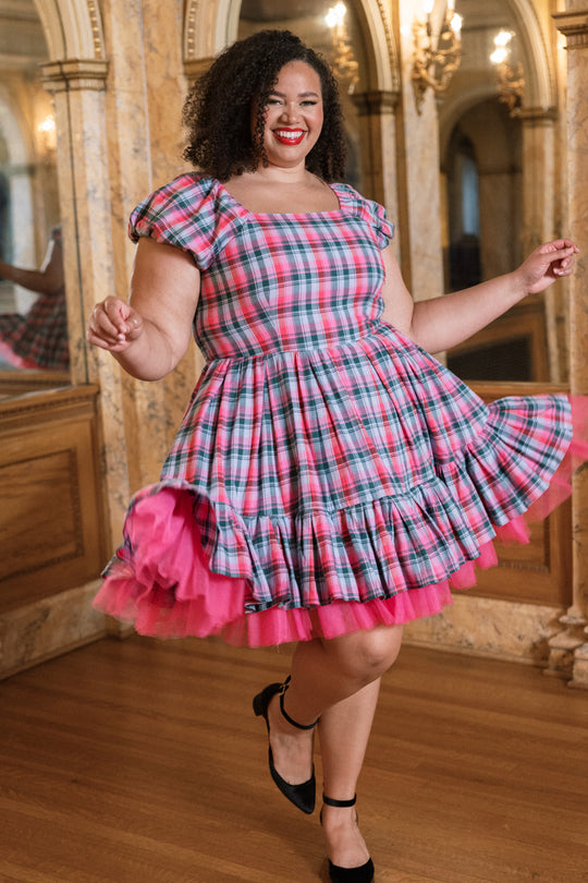 A woman beams and twirls in an elegant room adorned with ornate mirrors and wooden floors, showing off the playful charm of the Ivy City Co Scottie Dress in Vibrant Plaid.