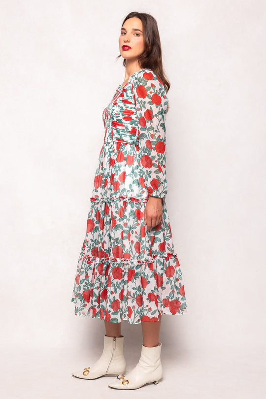 A woman stands sideways against a plain light background, wearing the Ivy City Co Selena Dress in Red Roses—a long, tiered dress with white and red floral print and long sleeves—paired with white ankle boots featuring gold accents; the flowing silhouette provides a chic, maternity-friendly style.