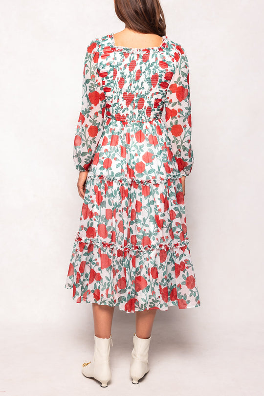 A woman stands facing away, wearing the Ivy City Co Selena Dress in Red Roses—a maternity-friendly, ankle-length dress featuring long sleeves and tiered layers adorned with vibrant red flowers and green leaves. She completes the look with white ankle boots.