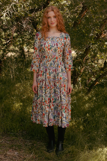 A woman with long curly red hair stands outdoors in a sunlit grassy area, wearing the Ivy City Co Selena Dress in Vivid Meadow and black boots, surrounded by trees and greenery.