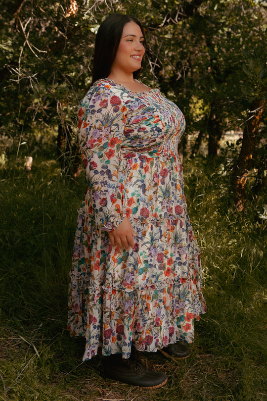 A woman stands sideways in a sunlit forest, smiling as she wears the Ivy City Co Selena Dress in Vivid Meadow—a floral midi with a smocked bodice and tiered ruffle skirt—paired with black boots among trees and grass.