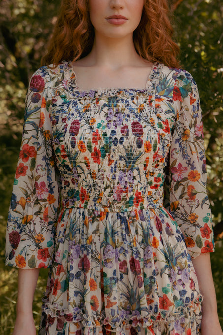 A woman with long, wavy red hair wears the Ivy City Co Selena Dress in Vivid Meadow, a floral midi with smocked bodice and puffed sleeves. She stands outdoors in natural sunlight, her face partly visible amid green foliage.