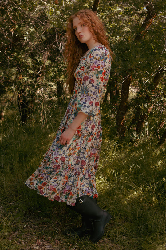A person with long, wavy red hair wears the Ivy City Co Selena Dress in Vivid Meadow with black boots, standing on grass in a sunlit forest surrounded by green trees and foliage.