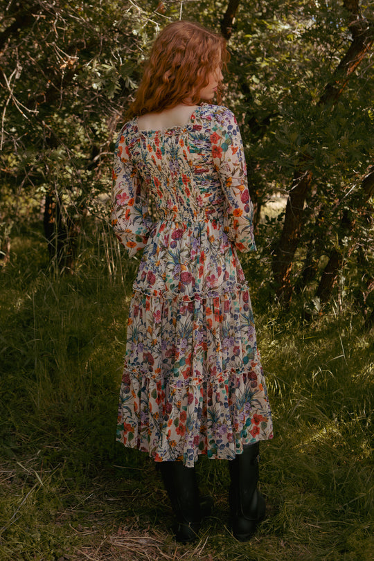 A person with wavy, reddish hair stands outdoors on grass, wearing the Ivy City Co Selena Dress in Vivid Meadow—a floral midi with a smocked bodice and tiered ruffle skirt—paired with black boots, surrounded by lush trees and greenery.