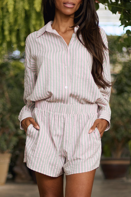 A woman wears Ivy City Co’s Sevilla Shorts in Green & Pink Pinstripe, paired with a matching long-sleeve, button-up shirt, standing outdoors with her hands in her pockets.