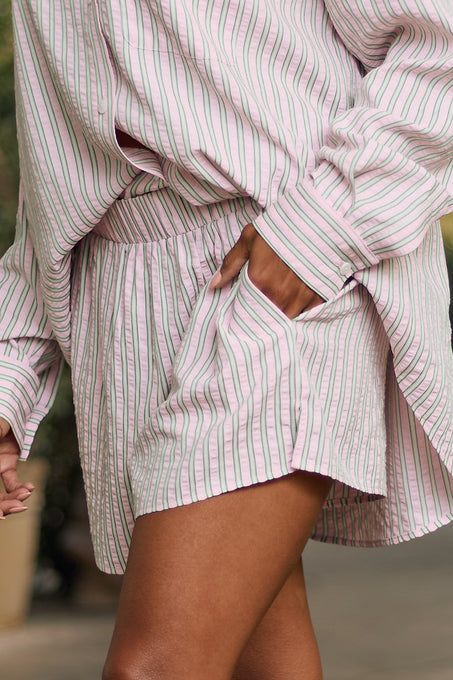A person wearing Ivy City Co’s Sevilla Shorts in Green & Pink Pinstripe and a loose-fitting, long-sleeve, pink and white striped shirt, with one hand in their pocket, stands outdoors.