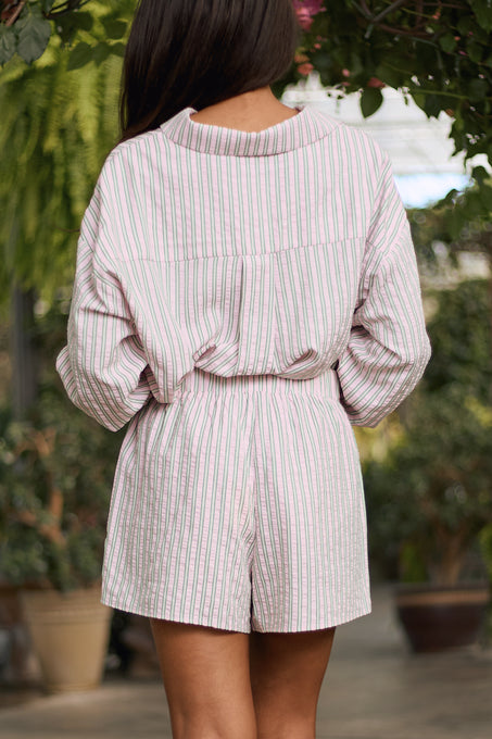 A person with long dark hair stands in a garden, surrounded by greenery, wearing Ivy City Co’s Sevilla Shorts in Green & Pink Pinstripe and a matching long-sleeve shirt, creating a fresh summer vibe.