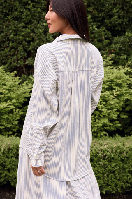 A woman with long dark hair stands outdoors by green bushes and trees, wearing the Ivy City Co Sevilla Top in Blue & White Pinstripe, a loose-fitting, nursing friendly shirt paired with matching pants.