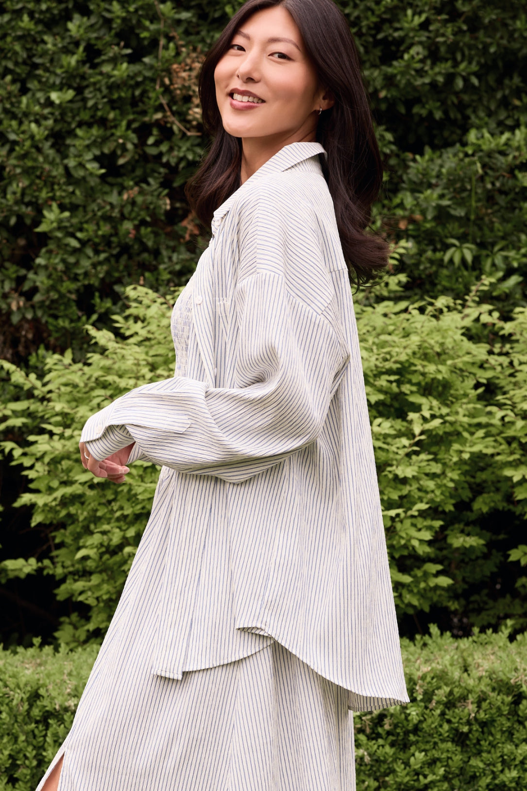 A woman with long dark hair smiles while walking outdoors in front of lush green bushes, wearing the Ivy City Co Sevilla Top in Blue & White Pinstripe.