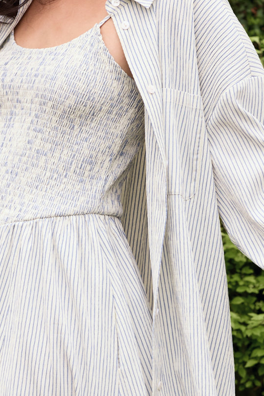 A person stands outdoors among greenery, wearing the Ivy City Co Sevilla Top in Blue & White Pinstripe—a light-colored top with vertical stripes and a relaxed, summery feel.