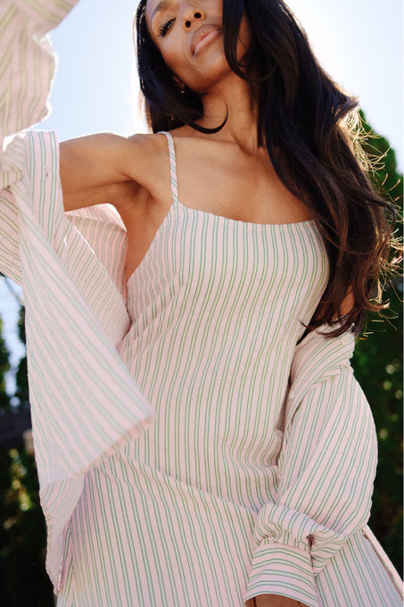 A woman with long, wavy hair wears the Ivy City Co Sevilla Top in Green & Pink Pinstripe. She stands outdoors in sunlight, looking confidently at the camera with greenery behind her.