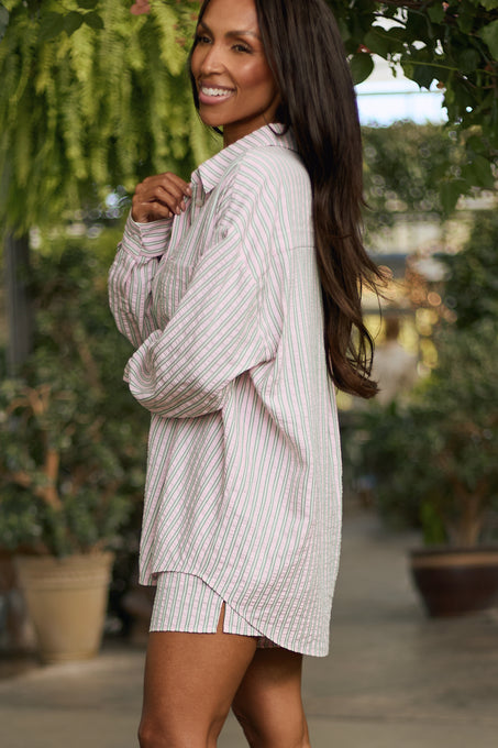 A woman with long dark hair smiles outdoors among potted plants, wearing the Ivy City Co Sevilla Top in Green & Pink Pinstripe—a nursing-friendly, loose-fit striped button-up with matching shorts—holding the shirt collar with one hand.