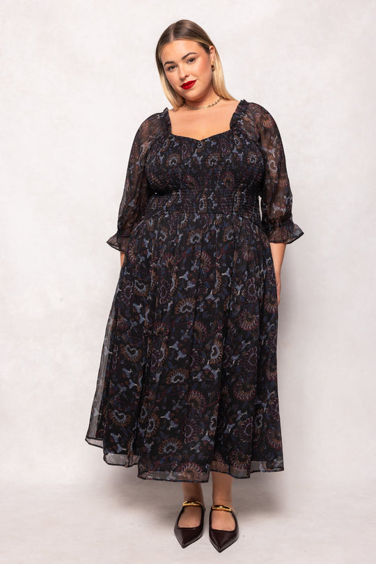 A woman wearing the Ivy City Co Maddison Dress in Enchanted Floral—a long, sheer black dress with puffed sleeves—stands against a softly textured backdrop. She has blonde hair, red lipstick, and is styled with brown pointed-toe flats. The ensemble exudes elegance and is also maternity friendly.