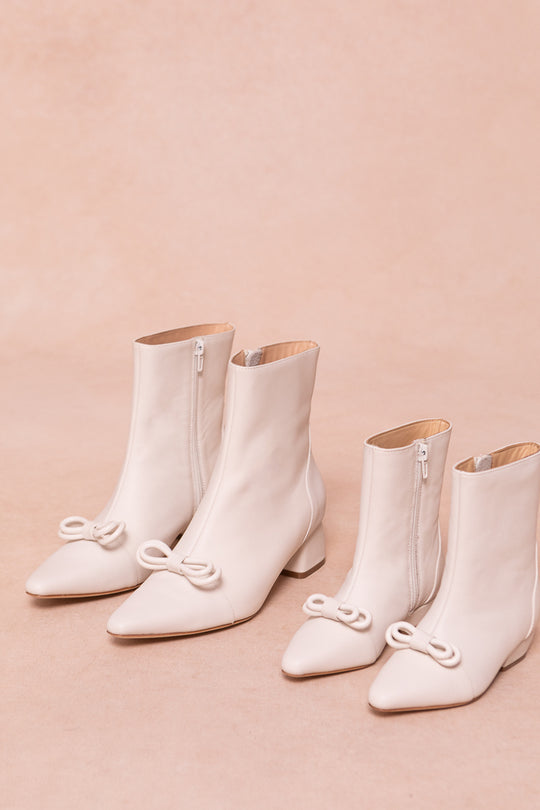 Two pairs of Ivy City Co’s Ivy Bow Ankle Boot in White, featuring matching cream color, side zippers, pointed toes, and decorative bows, are displayed on a light beige background. These chic boots add elegance to any look.