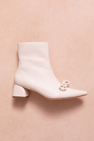 The Ivy Bow Ankle Boot in White by Ivy City Co features a pointed toe, low block heel, and a knotted detail on the upper—ideal for chic style lovers. Displayed against a light beige background.