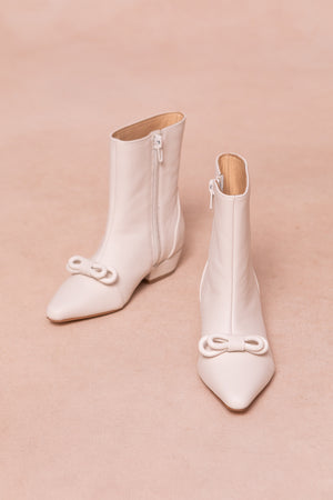 The Mini Ivy Bow Ankle Boot in White by Ivy City Co features white leather, pointed toes, side zippers, and decorative front bows, displayed on a light beige background.