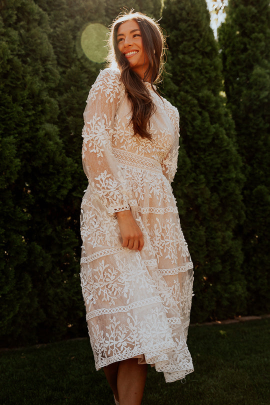 A woman smiles outdoors among tall green trees, wearing the Ivy City Co Sicily Dress with a champagne lining—long-sleeved and featuring a delicate white lace floral overlay.