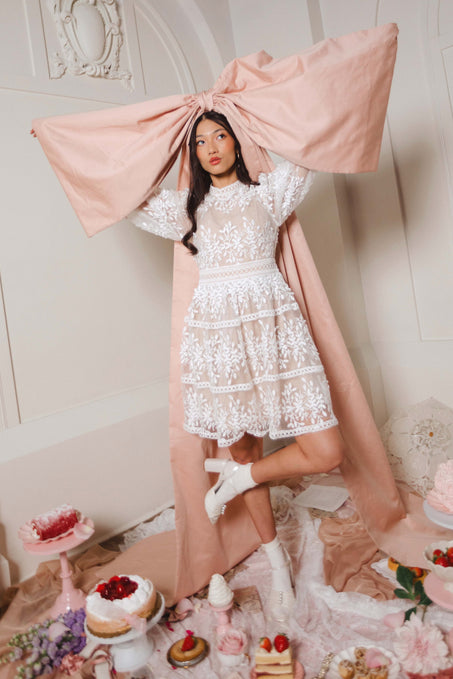 A woman wearing the Ivy City Co Sicily Short Dress and white boots stands with one leg raised, holding a large pink bow above her head. She is surrounded by cakes, pastries, and pink fabric in an ornate vintage-style room.