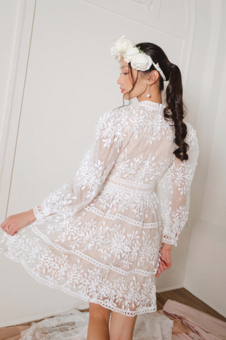 A woman wearing the Sicily Short Dress by Ivy City Co stands indoors on a light wooden floor, her back slightly turned. Her long dark hair is styled in a ponytail with white flowers.