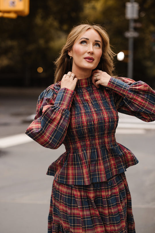 A woman with light brown hair, wearing the Ivy City Co Sigrid Top in Twilight Plaid—a long-sleeve red and blue plaid blouse with a mock neckline—stands outdoors near a street crosswalk, looking slightly upward with her hands touching her collar.