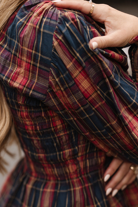 A close-up of a person wearing the Sigrid Top in Twilight Plaid by Ivy City Co, featuring red and navy plaid with balloon sleeves; one hand rests on their shoulder, highlighting manicured nails and a gold ring.