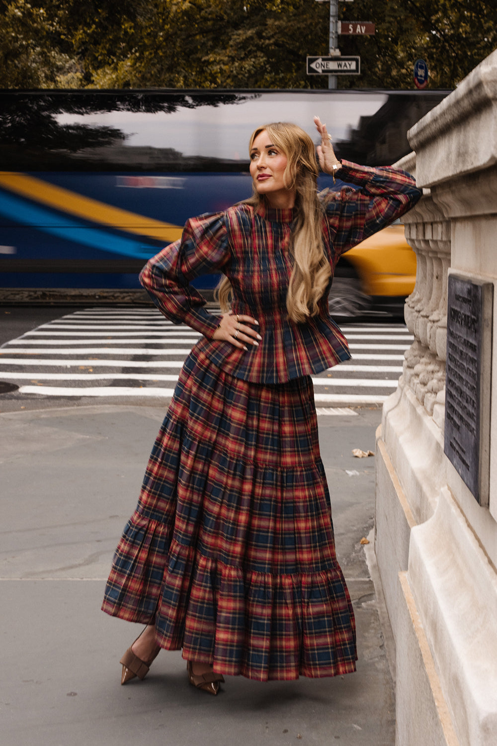 Woman wearing the Ivy City Co Sigrid Top in Twilight Plaid, featuring balloon sleeves and a mock neckline, poses by a stone railing on a city sidewalk with blurred traffic and trees in the background.