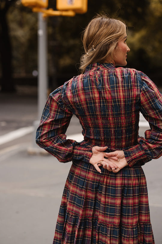 A woman wearing the Ivy City Co Sigrid Top in Twilight Plaid, featuring long balloon sleeves, stands outdoors with her back to the camera and hands clasped behind her. A blurry street and yellow traffic lights are visible in the background.