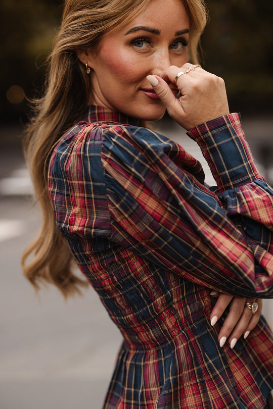 A woman with long hair poses outdoors in the Sigrid Top in Twilight Plaid by Ivy City Co, smiling softly with her hand near her face to show off manicured nails and rings.