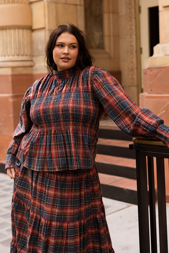 A woman wearing the Ivy City Co Sigrid Top in Twilight Plaid, featuring a mock neckline and long balloon sleeves, stands outdoors by a black railing with stone buildings and steps in the background.