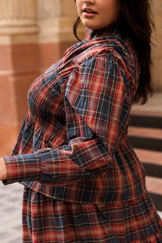A woman wearing the Ivy City Co Sigrid Top in Twilight Plaid, featuring a mock neckline and balloon sleeves, stands outdoors near steps, with her face partially visible.