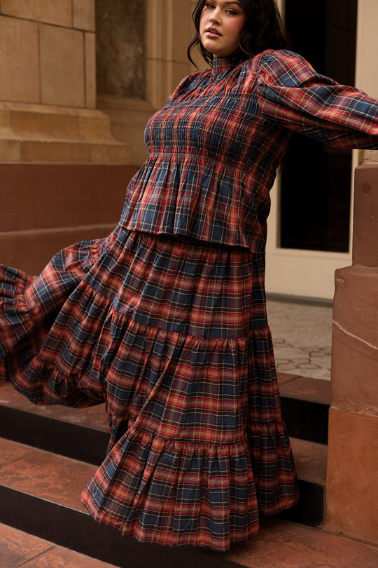 A woman wearing the Ivy City Co Sigrid Top in Twilight Plaid, featuring dramatic balloon sleeves and a long, tiered silhouette with red, navy, and beige tones, stands on outdoor steps and leans against a brown wall with one foot lifted.