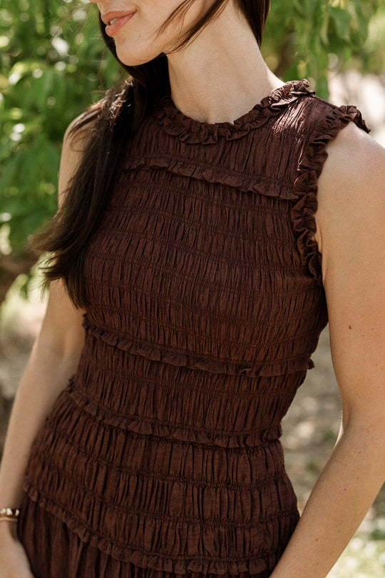A woman wearing the Simone Dress in Warm Cocoa by Ivy City Co—a sleeveless, textured brown dress with a smocked bodice—stands outdoors near green foliage, her long dark hair draped over one shoulder. Her face is partially visible as she smiles gently. This style is also maternity friendly.