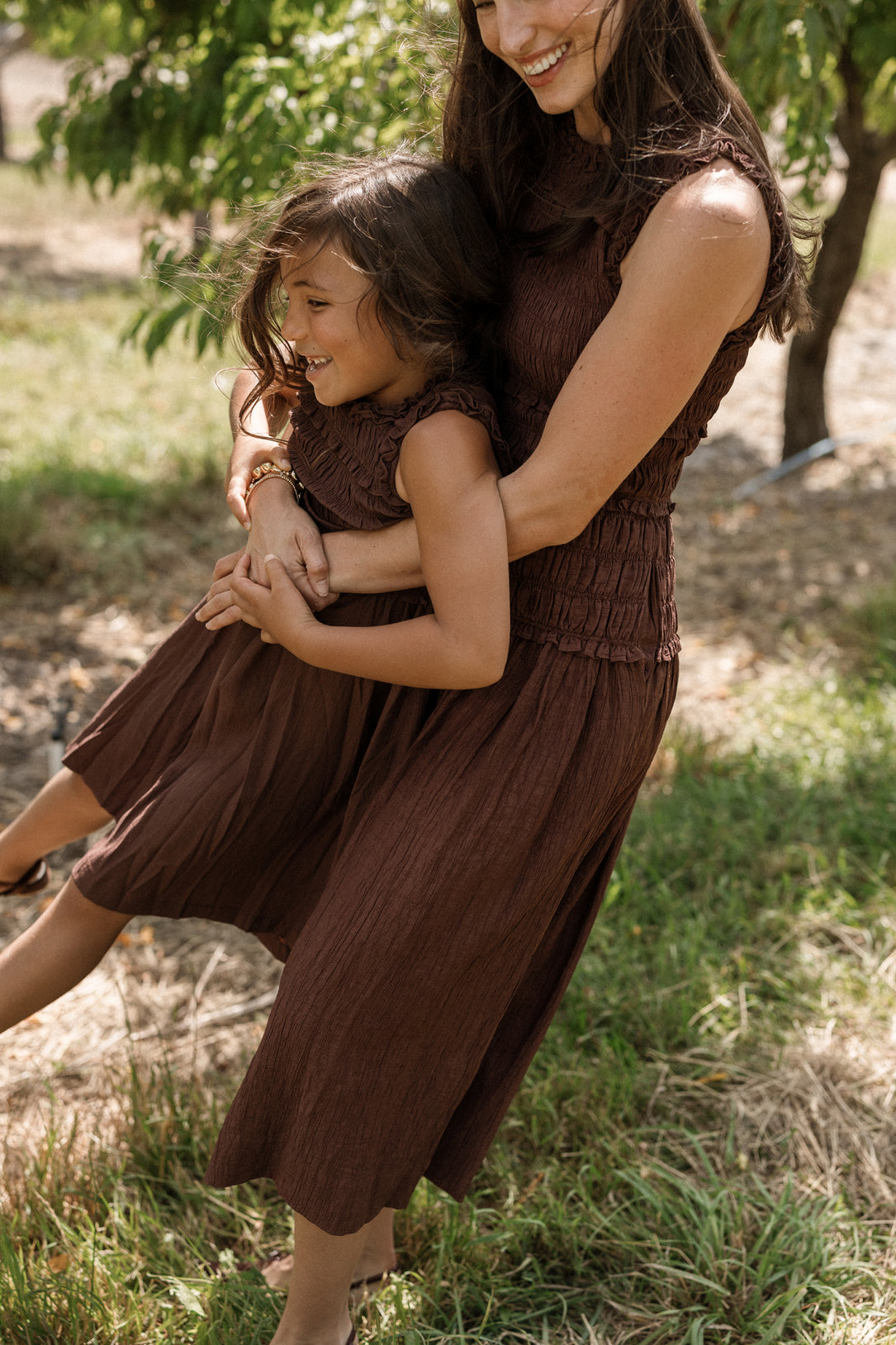 A woman and a young girl, both wearing matching Ivy City Co Simone Dresses in Warm Cocoa featuring a smocked bodice, smile and embrace while standing outdoors on a sunny day with green grass and trees in the background. The dresses are maternity friendly, making them perfect for every stage of life.