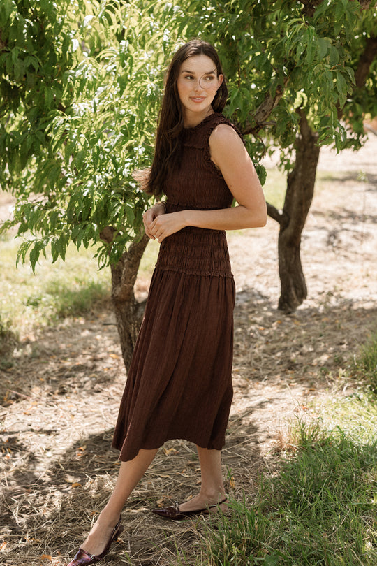 A woman stands outdoors among green trees, wearing the Ivy City Co Simone Dress in Warm Cocoa. She looks to the side with a slight smile as sunlight filters through the leaves, casting dappled shadows on the ground. The dress features a flattering, maternity-friendly smocked bodice and a sleeveless design.